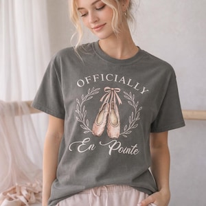 Op de afbeelding: Donkergrijs T-shirt met de tekst "OFFICIALLY En Pointe" en een afbeelding van balletschoenen. Het ontwerp is omgeven door een krans. Het model draagt een roze joggingbroek.