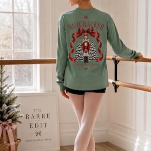 May include: A sage green long-sleeve shirt featuring a graphic on the back of a nutcracker soldier standing between two snow-covered pine trees, framed by a red ribbon and the text NUTCRACKER in red lettering above the design.