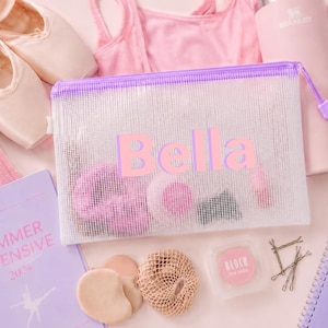 May include: A rectangular white mesh zipper pouch with a purple trim and the name Bella printed in large pink letters on the front. The translucent pouch contains various dance accessories, including pink toe pads and hair items, visible through the mesh.