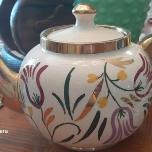 May include: A white ceramic teapot with gold trim and handle, featuring floral designs in red, yellow, and green. The teapot has a lid with a gold knob and is likely vintage.