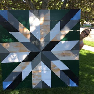 May include: Large, geometric wooden wall art featuring a starburst design. The artwork incorporates various shades of green, gray, black, and white. The piece is displayed outdoors, leaning against a grassy area.