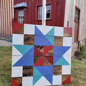 Puede incluir: Una colcha de granero de madera con un patrón de estrella en rojo, azul, blanco y turquesa. La colcha está hecha de tablones de madera recuperados y está colgada en el lado de un granero.