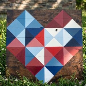Puede incluir: Arte de pared de madera en forma de corazón con un diseño geométrico. El corazón está compuesto por triángulos pintados en tonos de rojo, blanco y azul, sobre un fondo de madera marrón oscuro. La obra de arte se exhibe al aire libre en una zona de césped.