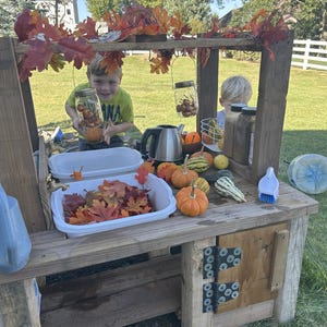 May include: A wooden play kitchen set outdoors, decorated with fall leaves. The set includes a sink, counter space, and a small cabinet. Various gourds and pumpkins are arranged on the counter. Two children are playing with the set.