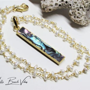 May include: A gold-toned necklace with a rectangular pendant featuring iridescent abalone shell. The necklace has two strands of small, white pearls and a gold clasp. The words "Hello Beach Vibes" are written in cursive at the bottom.