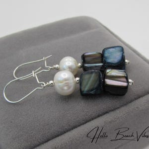 May include: A pair of dangle earrings with white pearls and iridescent square beads. The earrings have silver-colored hooks and are displayed on a gray velvet surface. The text "Hello Beach Vibes" is visible.