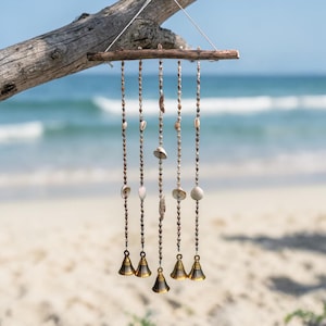 May include: A decorative wind chime made of natural materials. The chime features strands of seashells and beads suspended from a wooden branch. Small brass bells hang at the bottom of each strand. The background shows a beach and ocean.