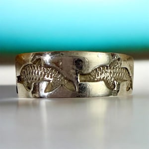 May include: A silver ring with a repeating pattern of two dolphins. The ring has a wide band and a polished finish. The dolphins are detailed with fins and curved bodies, creating a nautical theme.