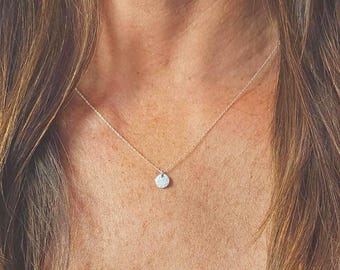 May include: A delicate silver-toned necklace featuring a small, circular pendant suspended from a thin, fine chain. The pendant has a smooth, reflective surface that catches the light, resting centrally against the collarbone area.