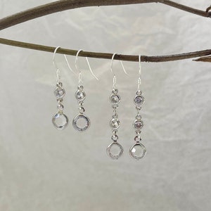 May include: Three pairs of silver dangle earrings with clear crystal stones. The earrings are hanging from a branch with dried pink roses.