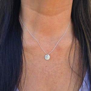May include: A delicate silver-toned necklace featuring a thin, fine chain and a small, circular pendant with a textured, hammered surface. The pendant hangs centrally from the chain, resting against the base of the neck in a simple, clean design.