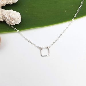 May include: A delicate silver-toned necklace featuring a thin, fine chain and a small, simple square-shaped pendant. The metal links of the chain are clearly visible, connecting to the minimalist square charm at the center of the necklace.