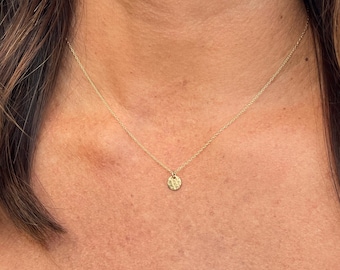 Peut inclure: Un collier doré délicat composé d'une fine chaîne et d'un petit pendentif circulaire à la surface martelée et texturée. Le pendentif est suspendu au centre de la chaîne, qui repose au niveau de la clavicule dans un design simple et épuré.