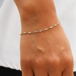 May include: Delicate gold and silver chain bracelet with small, rectangular links.  A hammered silver ring with an oval face complements the bracelet. Both pieces showcase a minimalist, modern aesthetic suitable for everyday wear. The jewelry is displayed on a wrist, highlighting its subtle elegance and refined design. The metal appears smooth and polished, reflecting light softly.