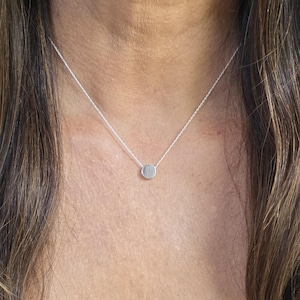 May include: A delicate silver-toned necklace featuring a small, circular pendant suspended from a thin, polished chain. The minimalist design centers on the smooth, round metal disc, which rests against the collarbone area on a fine link chain.