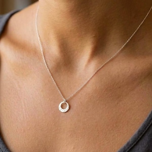 May include: A delicate silver necklace featuring a small, circular pendant with a textured, hammered finish. The pendant hangs from a thin, polished silver chain that rests against the collarbone, creating a simple and refined look.