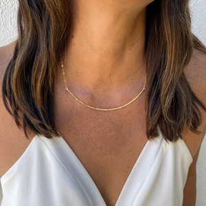 May include: A gold necklace with a thin, curved bar pendant. The necklace is worn by a person with long brown hair.