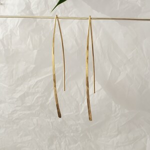 Pode incluir: Um par de brincos dourados longos e finos com um design em forma de V e um acabamento martelado e texturizado. Os brincos est&atilde;o suspensos num fio horizontal fino, exibindo a sua forma met&aacute;lica alongada, curva e superf&iacute;cie dourada.