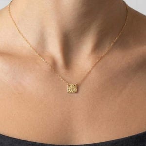 May include: A gold-toned necklace featuring a delicate chain and a small, square-shaped pendant with a textured, hammered surface. The pendant hangs centrally from the chain, which rests against the collarbone area.