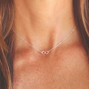 May include: A delicate silver-toned necklace featuring a small, centred infinity symbol pendant. The chain is thin and metallic, resting against the collarbone area, showcasing a minimalist design with a polished finish and a classic link structure.