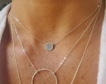 Könnte beinhalten: Eine silberne Halskette mit einem runden Anhänger und einer zarten Kette. Die Halskette ist mit zwei weiteren Ketten übereinander gelegt, eine mit einem Kreisanhänger und eine mit einer einfachen Kette.