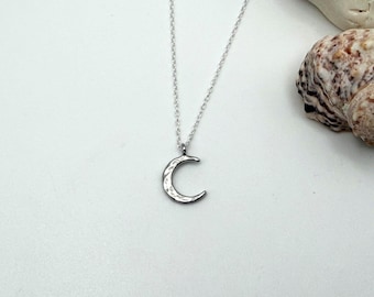 May include: A silver-toned necklace featuring a crescent moon pendant with a textured, hammered surface. The pendant hangs from a delicate, thin silver-toned chain. The metal has a reflective finish that catches the light, creating a subtle pattern.