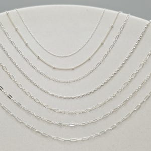 May include: A collection of delicate silver necklaces displayed on a white surface. The necklaces feature various chain styles, including beaded, cable, and figaro, showcasing a range of designs and textures. The chains are all silver in color.