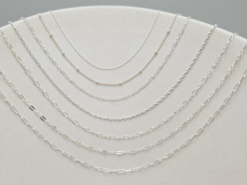 May include: A collection of seven silver-toned metal chains arranged in a cascading arc. The assortment includes various link styles, such as delicate cable chains, chains with small evenly spaced beads, and elongated oval link designs.
