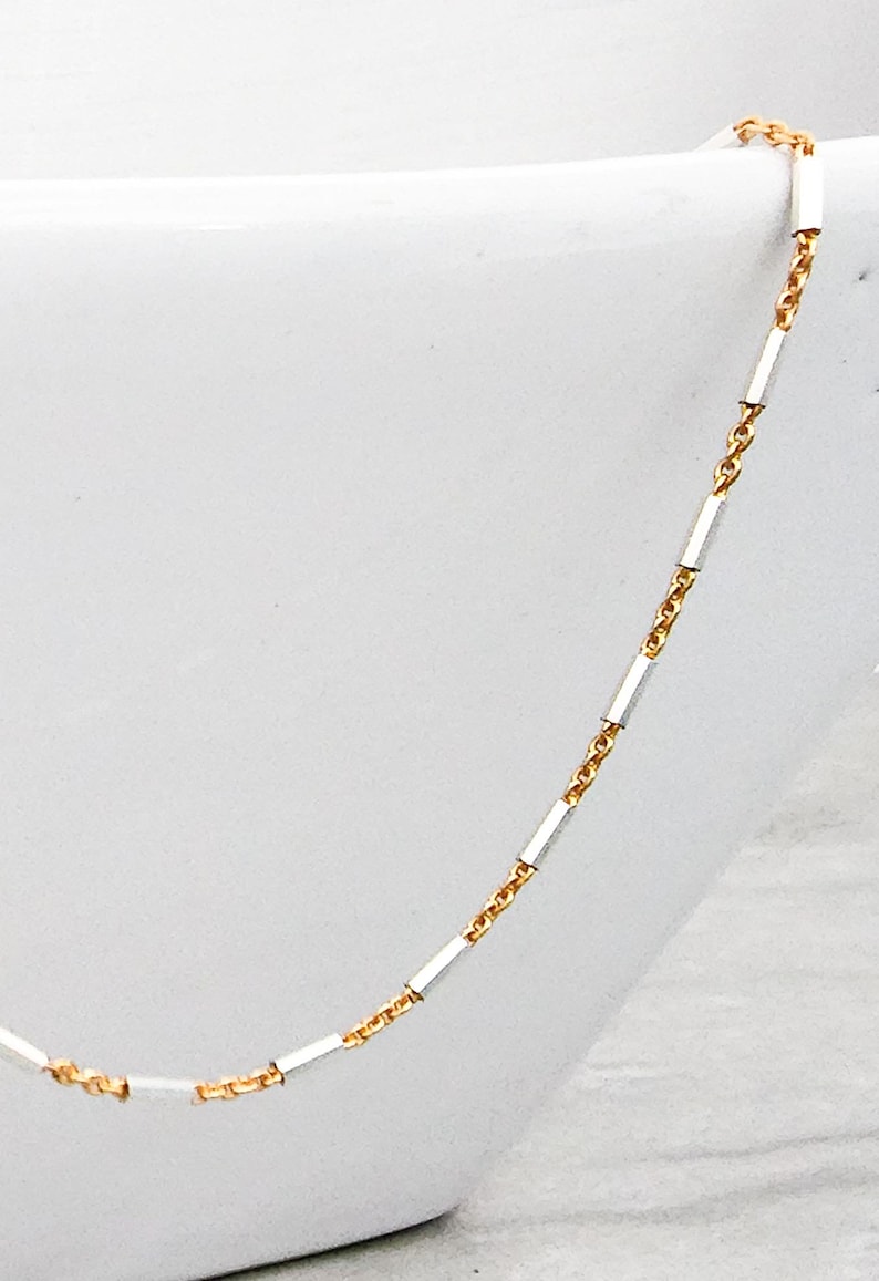 May include: A delicate gold chain necklace with silver bar accents. The chain is thin and has a delicate, feminine look.