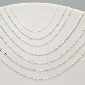 May include: A collection of eight distinct silver-toned metal chains arranged in a cascading arc. The assortment includes various link styles such as delicate cable chains, satellite chains with small beads, and elongated oval link chains.