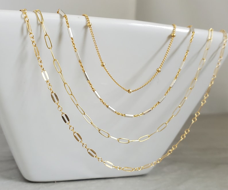 May include: Four gold chains of different lengths and styles, layered on a white surface. The chains are made of small, linked pieces of metal.