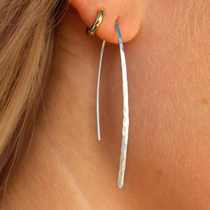 Pode incluir: Um pequeno brinco de argola em tom dourado e um brinco longo e fino de fio em tom prateado com um acabamento martelado e texturizado. O brinco prateado apresenta um design de barra alongada e curva que pende verticalmente.