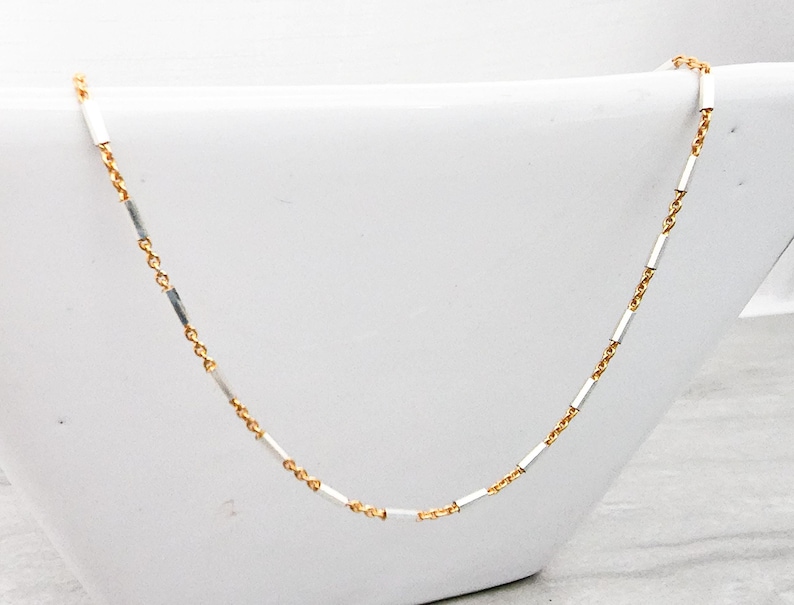 May include: A delicate gold and silver chain necklace with alternating square links. The chain is displayed on a white surface.