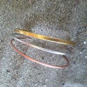 May include: Three metal bangles stacked on top of each other. The top bangle is gold, the middle bangle is silver, and the bottom bangle is copper.