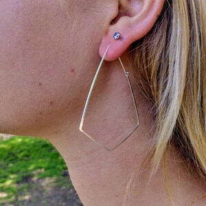 May include: Silver geometric hoop earrings with a diamond shape. The earrings are worn by a person with blonde hair.
