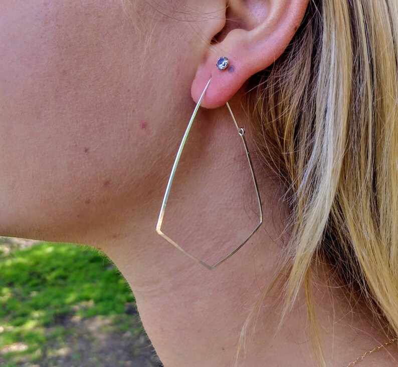 May include: Silver geometric hoop earrings with a diamond shape. The earrings are worn by a person with blonde hair.