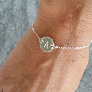 Könnte beinhalten: Ein silbernes Kettenarmband mit einem runden Anhänger, auf dem der Buchstabe 'A' eingraviert ist.