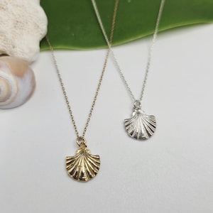 Puede incluir: Dos collares delicados con pequeños colgantes de concha marina texturizados. Un colgante tiene un acabado en tono dorado cálido, mientras que el otro tiene un acabado en tono plateado brillante. Cada colgante cuelga de una fina cadena de metal.