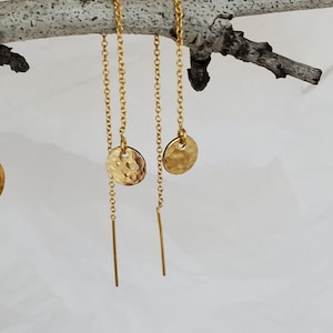 May include: Gold-colored chain earrings with hammered disc charms and long bar pendants. The earrings are hanging from a light-colored branch against a white background. The earrings are a delicate and elegant accessory.