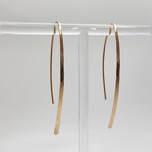 Pode incluir: Um par de brincos longos e curvos em tom dourado com um acabamento martelado e texturizado. Os brincos apresentam um design fino em forma de U alongado que pende verticalmente, exibindo um brilho met&aacute;lico e uma superf&iacute;cie facetada.