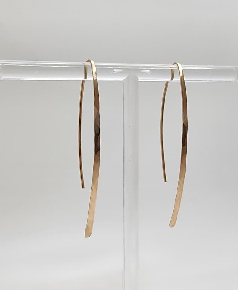 Pode incluir: Um par de brincos longos e curvos em tom dourado com um acabamento martelado e texturizado. Os brincos apresentam um design fino em forma de U alongado que pende verticalmente, exibindo um brilho met&aacute;lico e uma superf&iacute;cie facetada.