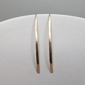 Pode incluir: Um par de brincos longos e finos de cor dourada com uma forma vertical curva. Cada brinco apresenta uma superf&iacute;cie martelada e texturizada que capta a luz, criando uma s&eacute;rie de pequenas facetas planas ao longo da barra de metal.