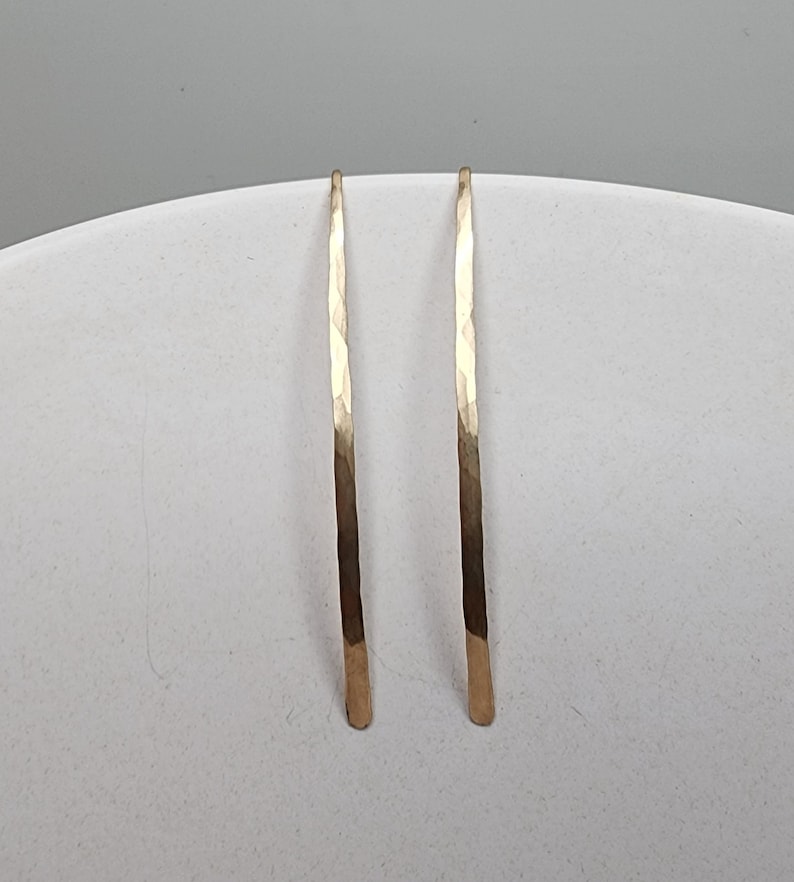 Pode incluir: Um par de brincos longos e finos de cor dourada com uma forma vertical curva. Cada brinco apresenta uma superf&iacute;cie martelada e texturizada que capta a luz, criando uma s&eacute;rie de pequenas facetas planas ao longo da barra de metal.