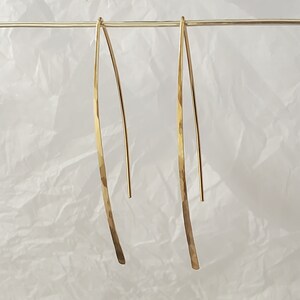 Pode incluir: Um par de brincos dourados longos e curvos com um acabamento martelado e texturizado. Os brincos est&atilde;o suspensos num fio horizontal dourado fino, exibindo um design elegante em forma de V alongado com uma superf&iacute;cie met&aacute;lica polida.