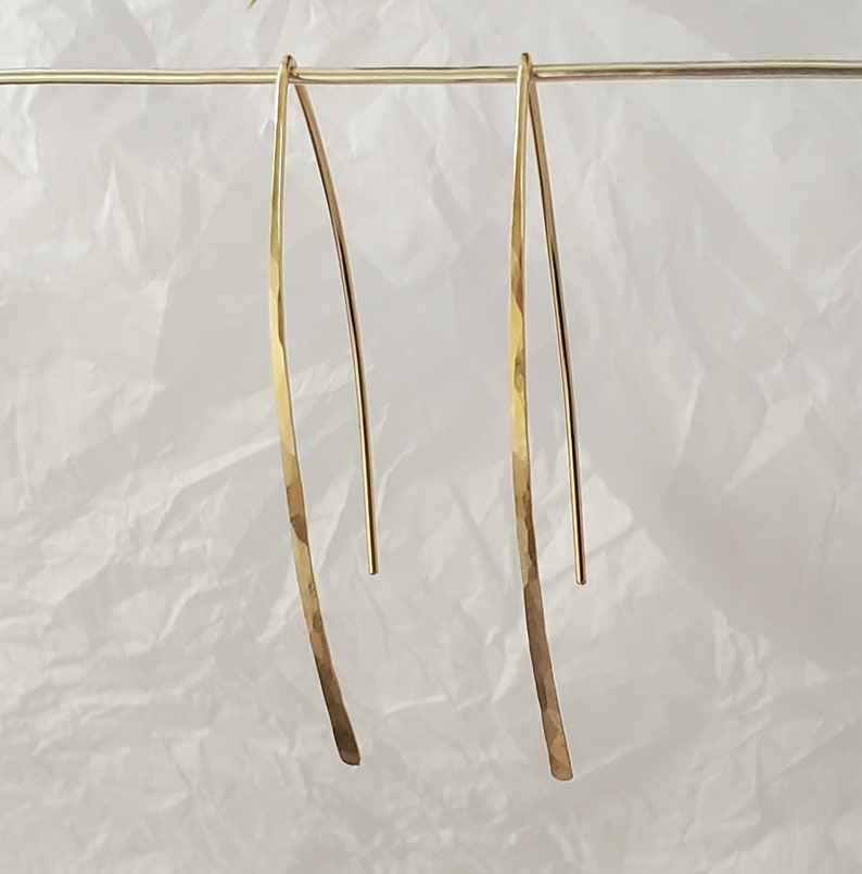 Pode incluir: Um par de brincos dourados longos e curvos com um acabamento martelado e texturizado. Os brincos est&atilde;o suspensos num fio horizontal dourado fino, exibindo um design elegante em forma de V alongado com uma superf&iacute;cie met&aacute;lica polida.