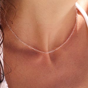 May include: A delicate silver-toned chain necklace featuring a series of elongated, rectangular links. The necklace rests against the skin, showcasing a simple, uniform design with a polished metallic finish that catches the light.