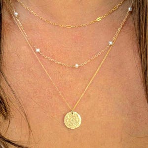 May include: A layered gold necklace set consisting of three distinct chains. The top chain features elongated links, the middle chain is adorned with small white pearls, and the bottom chain holds a round, textured gold pendant with a hammered finish.