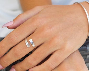 May include: A silver-colored wrap ring with a hammered texture is worn on a finger. Two silver bracelets are also visible on the wrist. The ring and bracelets are simple and elegant, suitable for everyday wear. The nails are painted with a light pink nail polish.