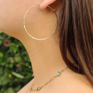 May include: Gold hoop earrings with a hammered texture. The earrings are worn by a person with long brown hair. The person is also wearing a gold necklace with iridescent blue stones.
