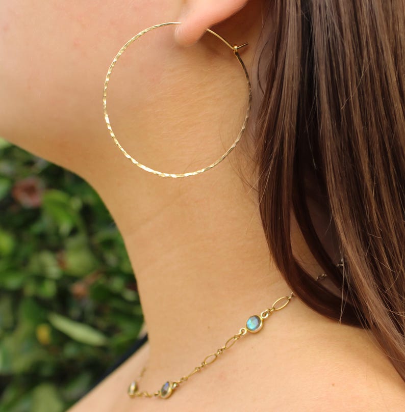 May include: Gold hoop earrings with a hammered texture. The earrings are worn by a person with long brown hair. The person is also wearing a gold necklace with iridescent blue stones.